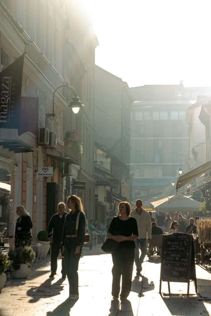 A sunlit cobblestone street in Sarajevo, bustling with people enjoying their one-day itinerary, is flanked by charming buildings and shops, including an inviting outdoor café. Signs and greenery adorn the scene, creating a lively urban atmosphere under a clear sky.