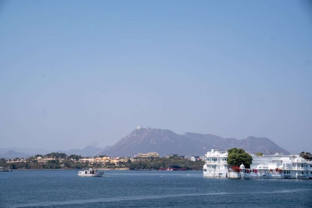 Scenic view of a lake with a boat floating on it, surrounded by distant hills. On the right, part of a 1-week Rajasthan itinerary, there is a white building with a red canopy, partially surrounded by trees and water. The sky is clear and blue.