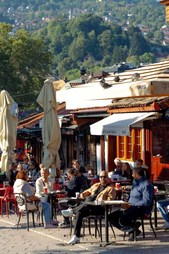 People sitting at outdoor café tables in a bustling street, perhaps enjoying the charms of a one-day in Sarajevo itinerary. Umbrellas are closed, pigeons rest on a nearby roof, while the backdrop reveals lush green hills adorned with scattered buildings under a clear sky.