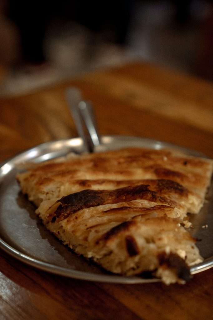 A slice of flaky, golden-brown pastry on a metal plate, placed on a wooden table, adds delight to your one-day Sarajevo itinerary. A fork rests beside the pastry, inviting you to savor each moment in this charming city.