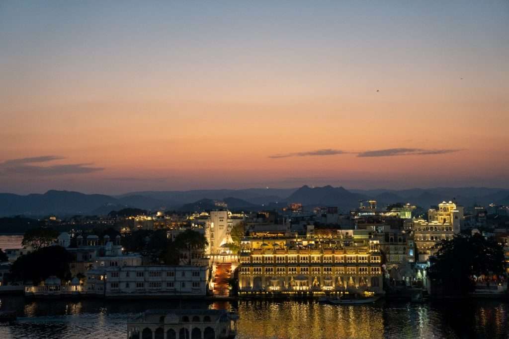 A scenic view of udaipur at dusk presents a softly lit cityscape reflecting on a tranquil lake, reminiscent of scenes from a 1-Week Rajasthan Itinerary. The sky's orange and blue hues cast an enchanting glow over the illuminated buildings, while distant hills line the horizon in peace.