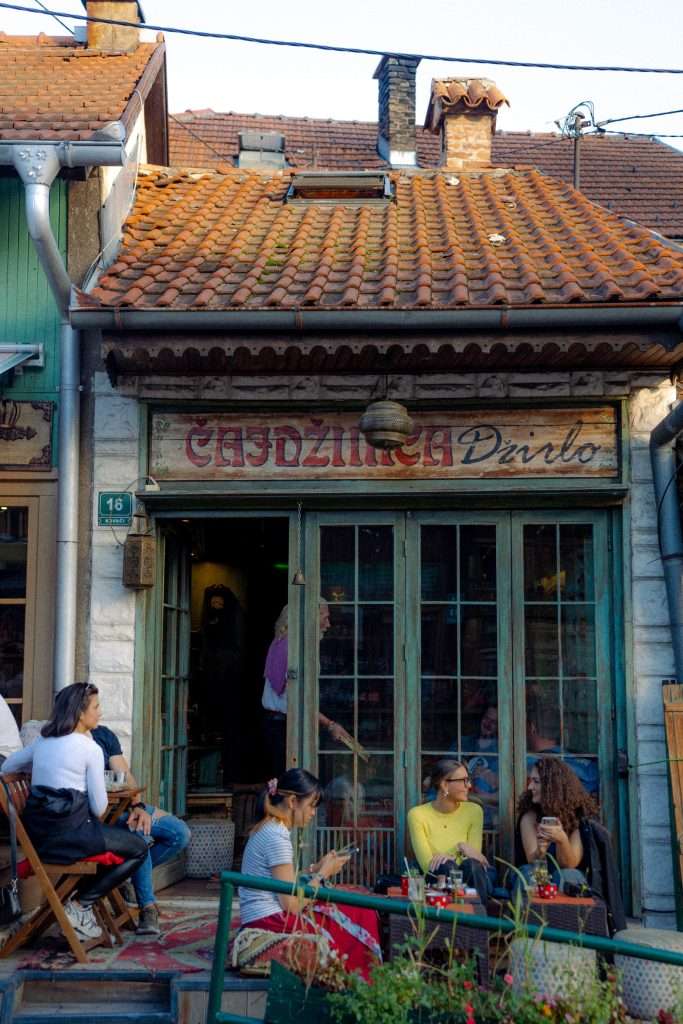 A group of people enjoys a leisurely chat outside a rustic café with a charming wooden facade, perfect for anyone crafting a one-day in Sarajevo itinerary. The cozy setting features chairs and a small table arranged beneath an old sign, inviting passersby to pause and savor the moment.