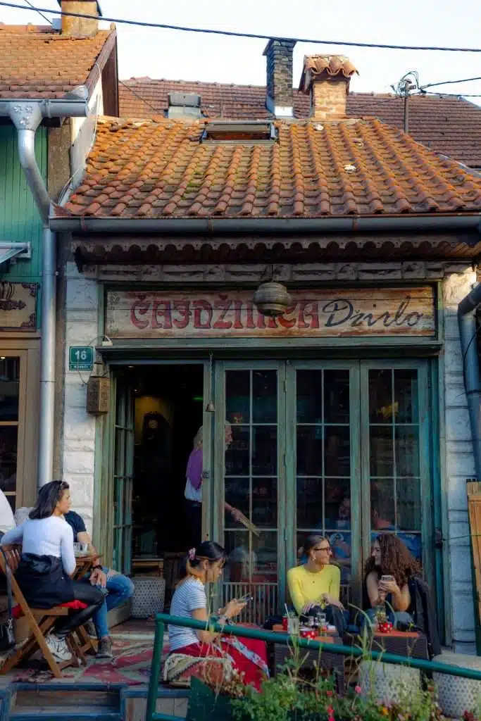 A group of people enjoys a leisurely chat outside a rustic café with a charming wooden facade, perfect for anyone crafting a one-day in Sarajevo itinerary. The cozy setting features chairs and a small table arranged beneath an old sign, inviting passersby to pause and savor the moment.