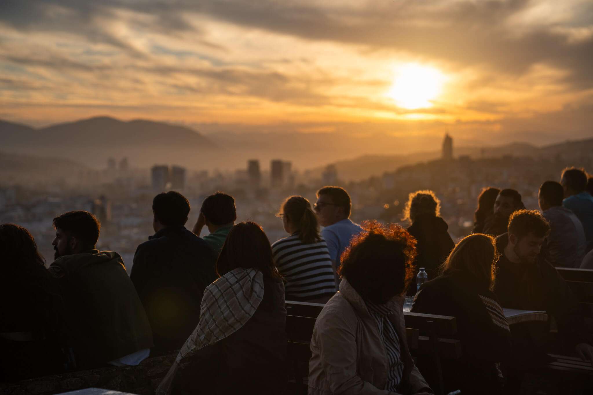 A group of people seated on benches enjoys a one-day in Sarajevo itinerary, overlooking a cityscape at sunset. The sun sinks behind rolling hills, casting a warm glow. Silhouettes of buildings and people create a serene, contemplative atmosphere.