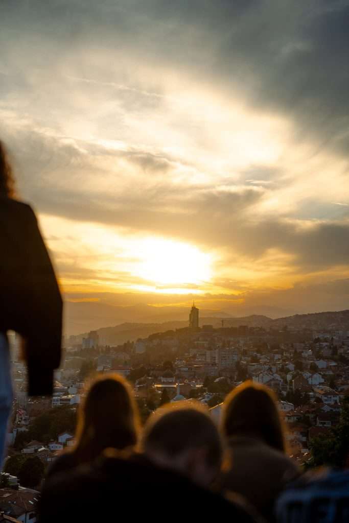 Silhouetted people overlook a cityscape during a golden sunset, reminiscent of the magical moments in a one-day Sarajevo itinerary. The sun casts a warm glow over the horizon, creating a dramatic sky with scattered clouds, while a prominent tower stands amidst the urban landscape - Yellow Fortress In Sarajevo