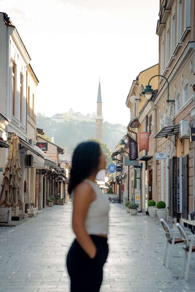 A woman stands on a cobblestone street lined with shops and cafes, absorbed in her one-day Sarajevo itinerary, as a distant mosque and hillside blur softly in the background. The street is quiet, basked in gentle morning or late afternoon light.