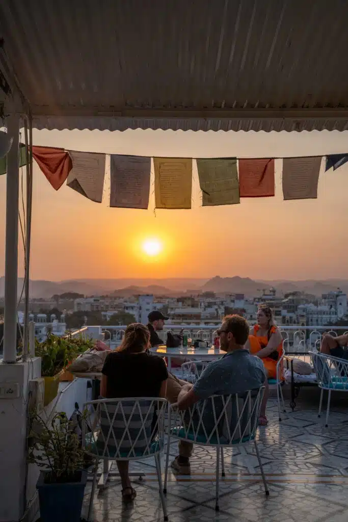 People relax at outdoor tables on a rooftop café at sunset, colorful prayer flags overhead, enjoying stunning views of the city and mountains—a perfect spot to pause during a 2-Week North India Itinerary.