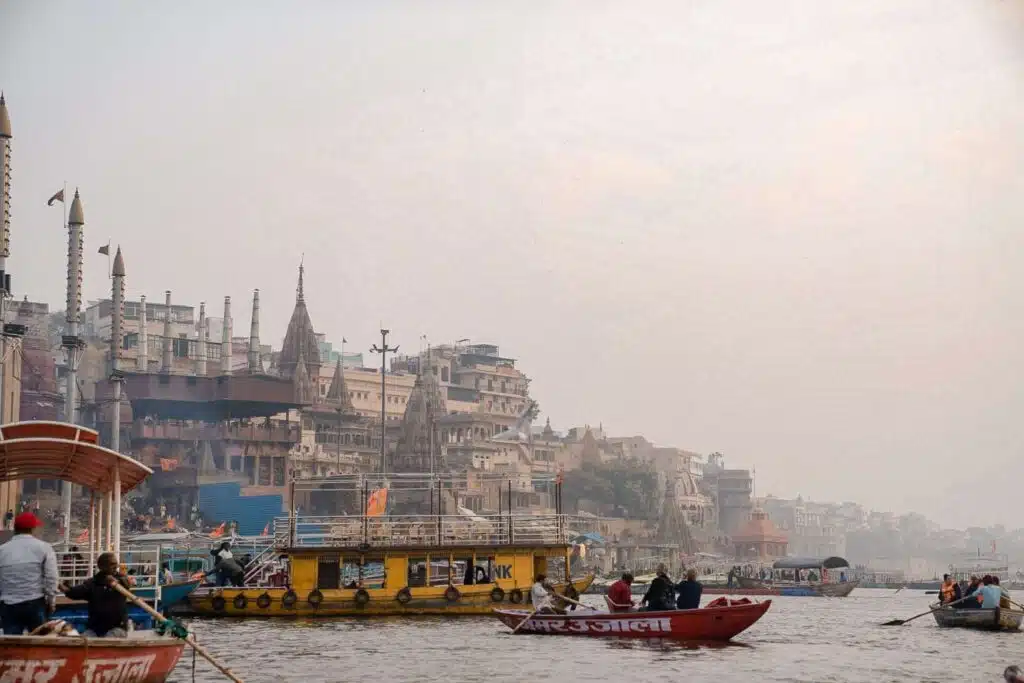 Boats with people float on the Ganges River near the crowded ghats of Varanasi, India—an essential stop on any 2-Week North India Itinerary—with historic buildings and temples lining the misty riverbank in the background.