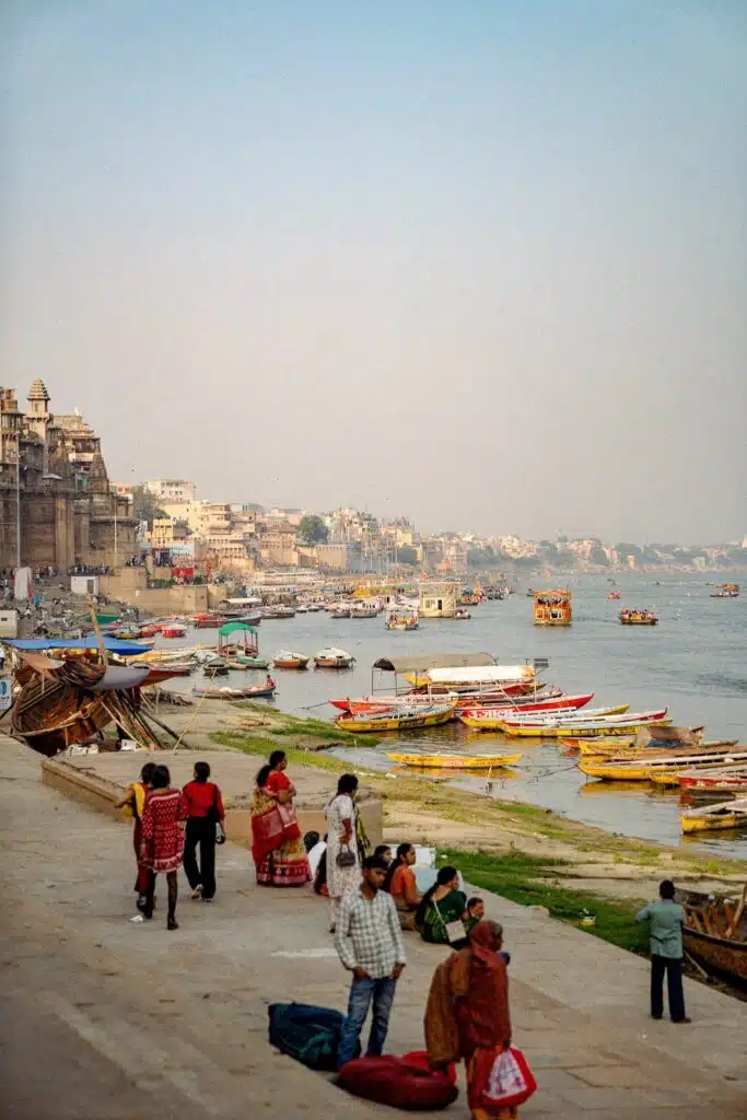 People stand and walk along the ghats of the Ganges River in Varanasi, India—an essential stop on any 2-Week North India Itinerary—with colorful boats docked and historic buildings lining the water under a clear sky.