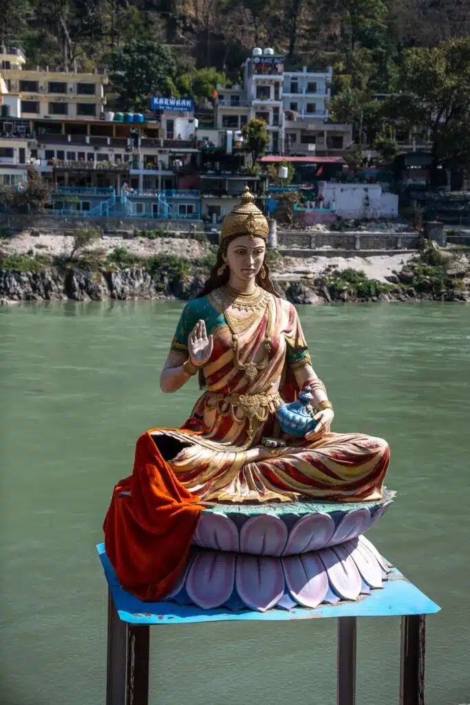 Backpacking India A colorful statue of a Hindu goddess sits cross-legged on a lotus pedestal beside a river, a serene scene you might encounter during your 2-Week North India Itinerary, with buildings and trees on the opposite bank.