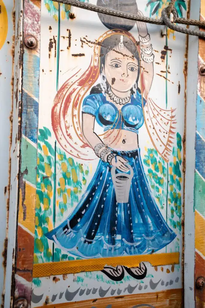 A colorful painting on a wooden surface depicts a woman in traditional Indian attire—perfect inspiration for your 2-Week North India Itinerary—as she balances a pot on her head amid vibrant green and yellow patterns.