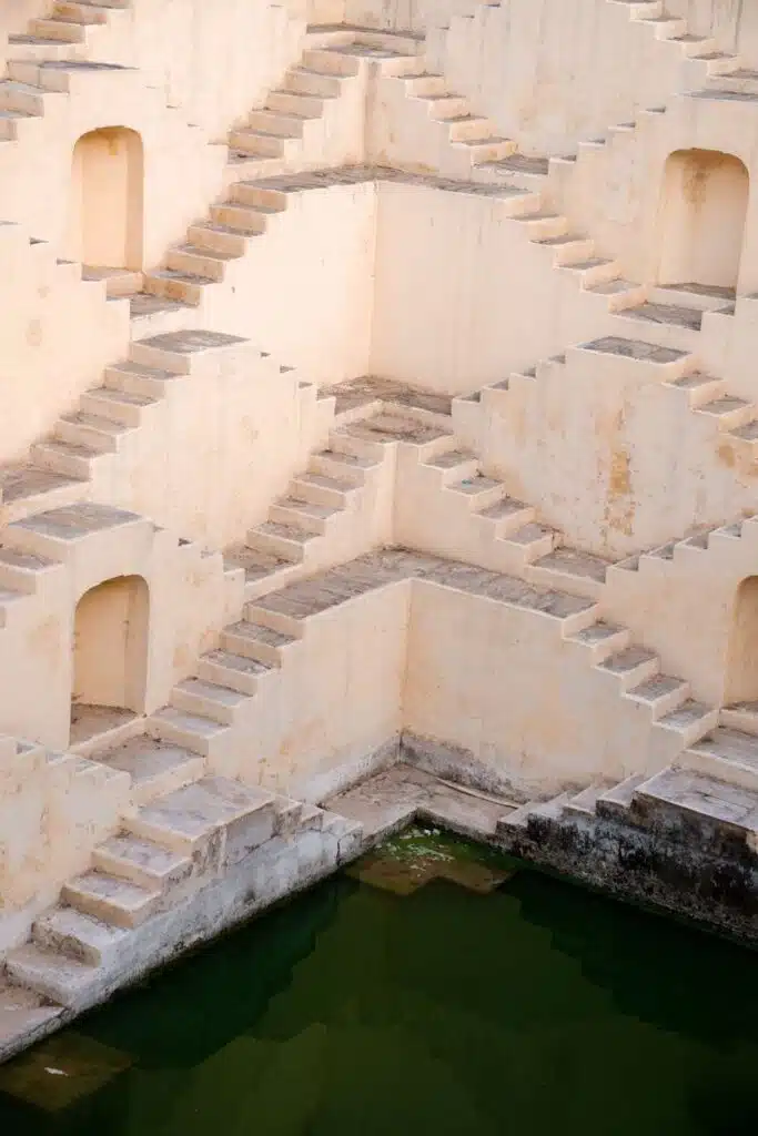 A highlight on a 2-Week North India Itinerary, this geometric stepwell features crisscrossing stone staircases descending to a pool of green water, bordered by pale, weathered walls and arch-shaped doorways.