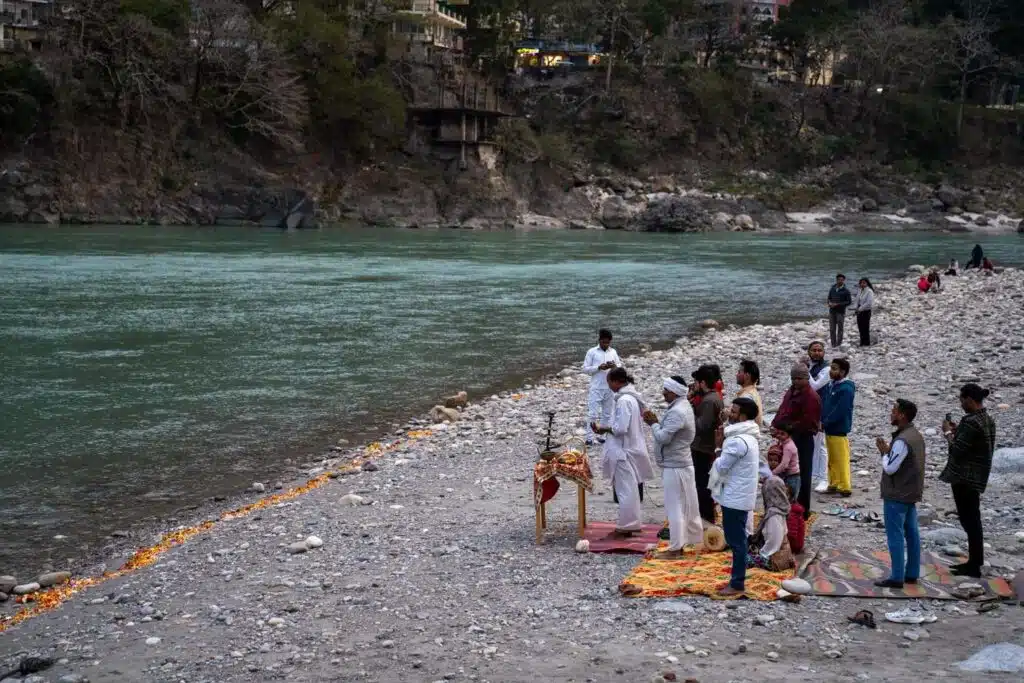 As part of a 2-Week North India Itinerary, a group stands and prays beside a river on a rocky shore. Some kneel on a colorful mat, led by individuals in white robes, while lit candles or lamps line the riverbank.