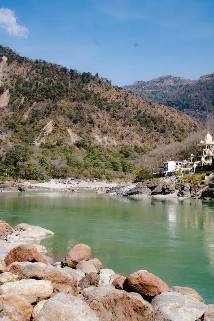 A calm green river flows past rocky banks and tree-covered hills under a clear blue sky, with a few buildings nestled on the hillside to the right—a tranquil scene often featured in a 2-Week North India Itinerary.
