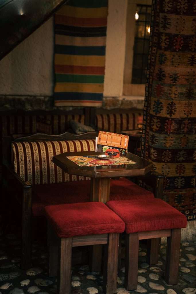 A cozy cafe setting in Cafe Divan Sarajevo perfect for your one-day in Sarajevo itinerary, features a small wooden table, two red cushioned stools, and striped pillows on a bench. Patterned rugs hang on the walls, enhancing the warm and inviting atmosphere.