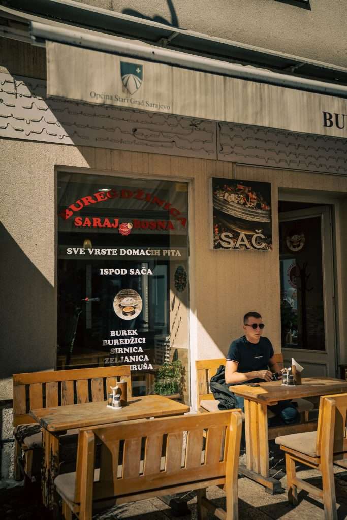 A person sits at an outdoor wooden table outside a bakery, basking in the sun's glow while planning their one-day in Sarajevo itinerary. Signage displays various pastries, adding to the cozy atmosphere as shadows dance around - Buregdžinica Saraj Bosna, Sarajevo