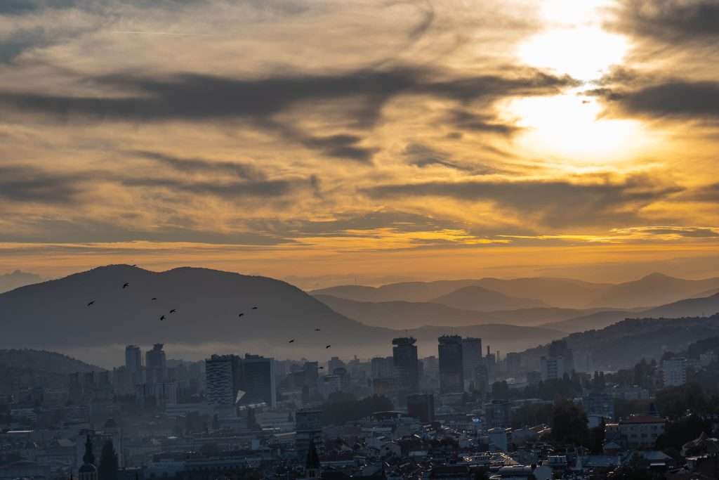 A panoramic view of Sarajevo's skyline at sunset reveals tall buildings silhouetted against the golden sky. Birds fly gracefully across the scene, while distant hills emerge under a dramatic cloud-streaked expanse—This is one of the best places to visit during 3 Days In Bosnia and Herzegovina 