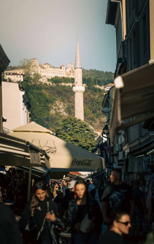 A bustling street scene unfolds in Sarajevo, with people strolling among market stalls. A tall minaret rises in the background against a hillside crowned with historic buildings, part of a perfect one-day itinerary. The sky is clear, and the lighting is warm.