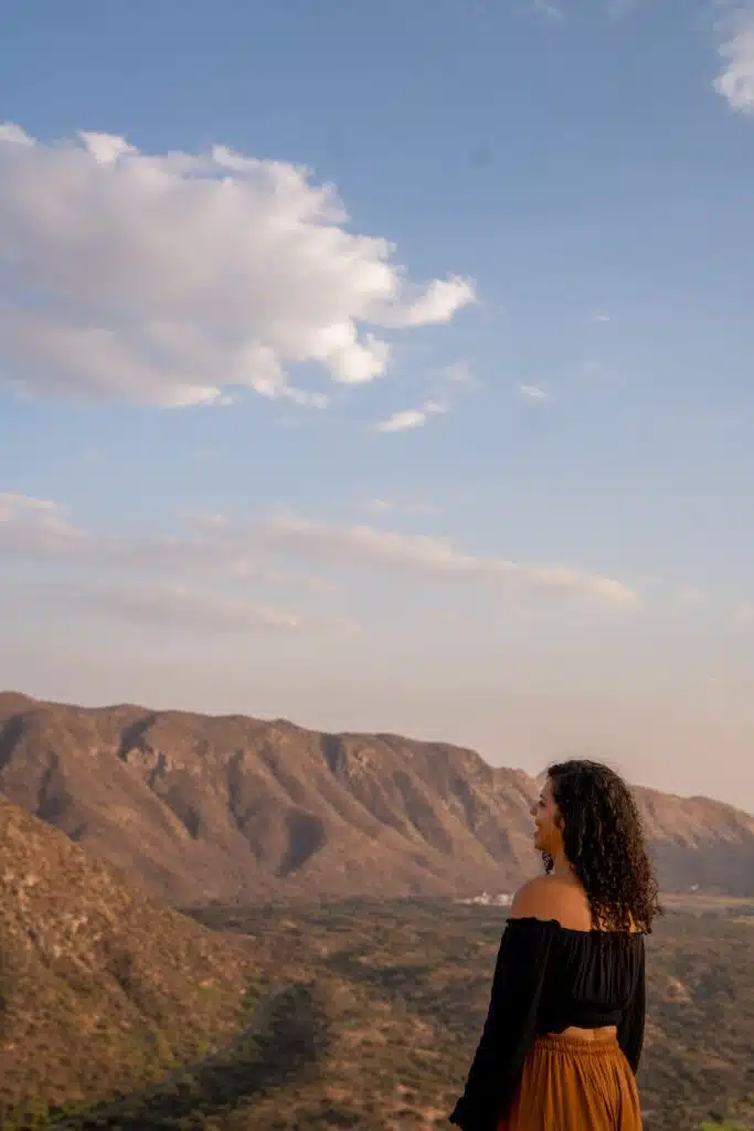 A woman with curly hair stands on a hillside, wearing a black off-shoulder top and a brown skirt, taking in the scenic mountain view—an inspiring moment for any 2-Week North India Itinerary.