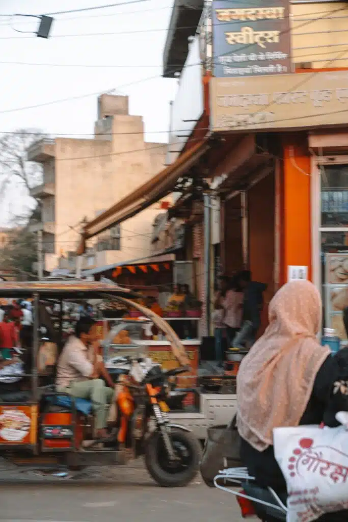 Backpacking India A busy street scene in India, perfect for a 2-Week North India Itinerary, with people riding a rickshaw and motorcycle. Buildings and shops line the street, while a woman in a headscarf carrying a bag stands in the foreground.