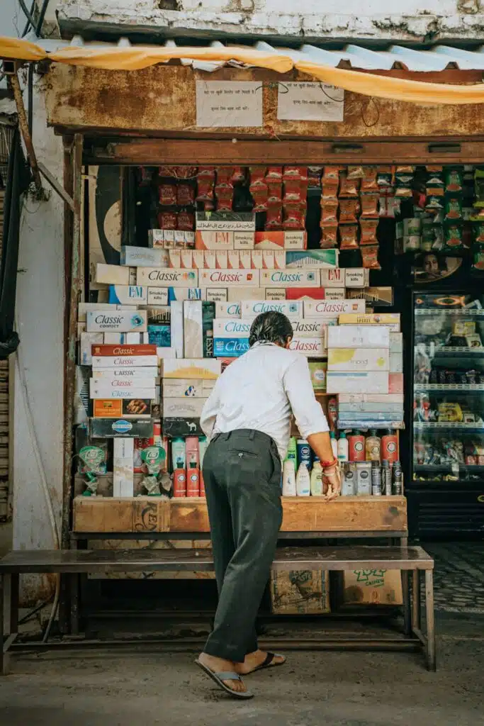 A man in a white shirt and dark pants, wearing flip-flops, leans over a wooden stall displaying boxes, drinks, and snacks—capturing the vibrant street life often found on a 2-Week North India Itinerary.