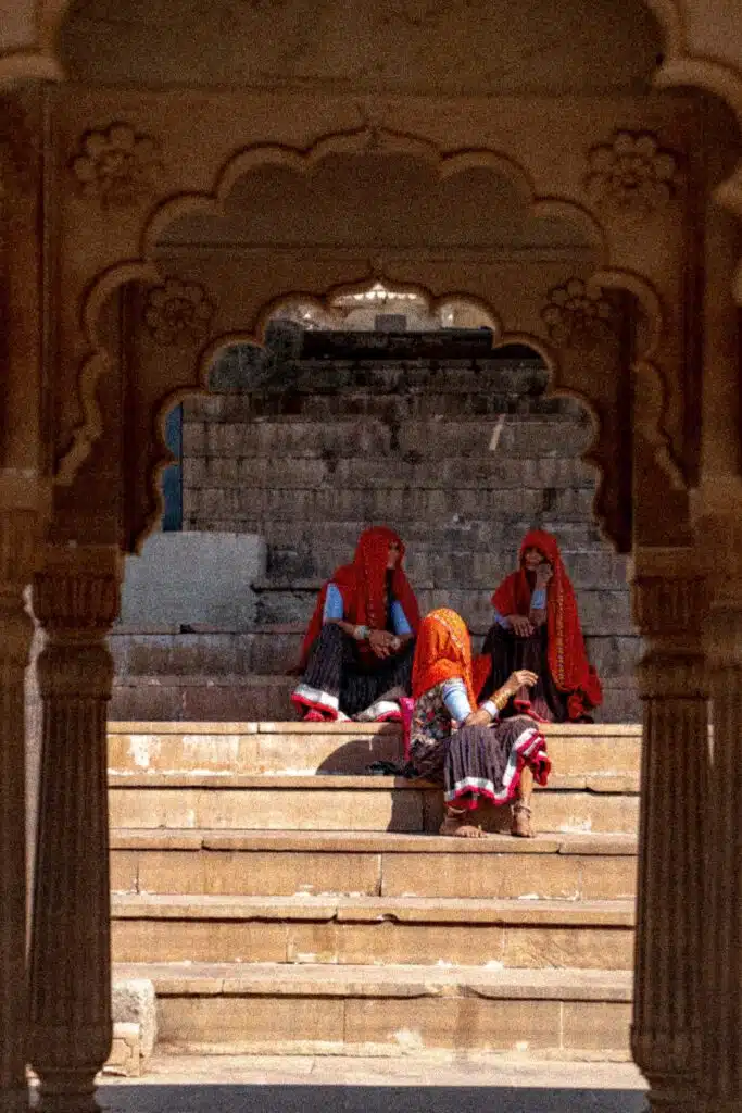 Three women in colorful traditional attire and red headscarves sit on stone steps, framed by an ornate arched doorway—an inviting scene you might encounter on a 2-Week North India Itinerary exploring ancient stone structures.