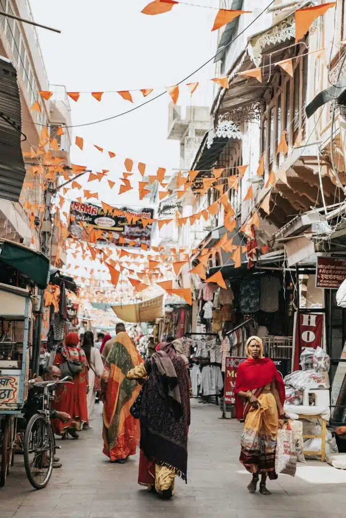 Backpacking India A lively street scene in India, perfect for a 2-Week North India Itinerary, shows women in colorful sarees walking through a market, with orange triangular flags overhead and shops lining both sides of the narrow pathway.