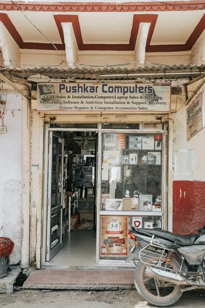 A small storefront named "Pushkar Computers" with a sign advertising computer sales, installation, repairs, and accessories—an unexpected stop along your 2-Week North India Itinerary. The open entrance reveals shelves of electronics beside a parked motorcycle.