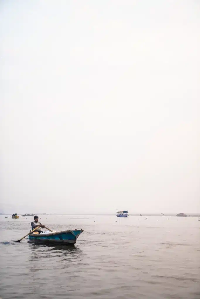 A person rows a small blue boat on a calm, wide river under a pale sky, with distant boats on the horizon—a tranquil scene reminiscent of peaceful moments from a 2-Week North India Itinerary.