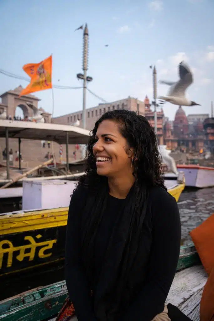 A smiling woman with curly hair sits in front of colorful boats on a river in India—an inspiring scene for your 2-Week North India Itinerary—with temples, flags, and a flying bird in the background under a partly cloudy sky.