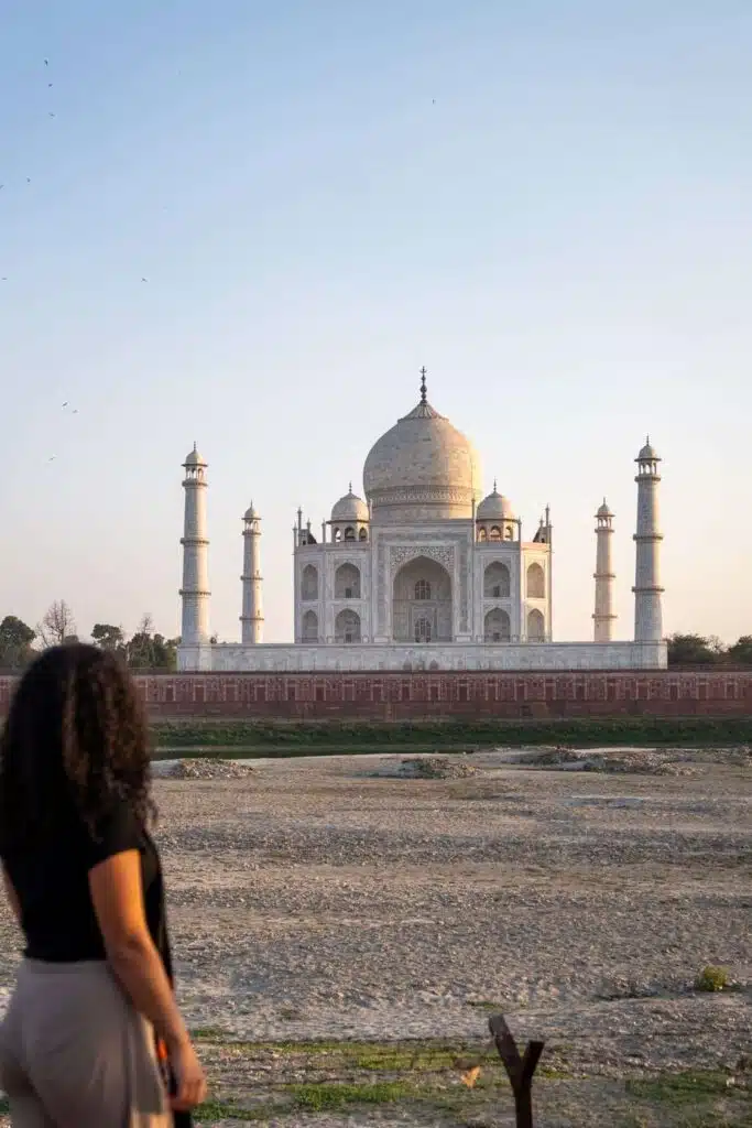 A woman with curly hair stands in the foreground, gazing at the white marble Taj Mahal under a clear sky—a highlight for any 2-Week North India Itinerary—while an open, dry area stretches between her and the iconic monument.