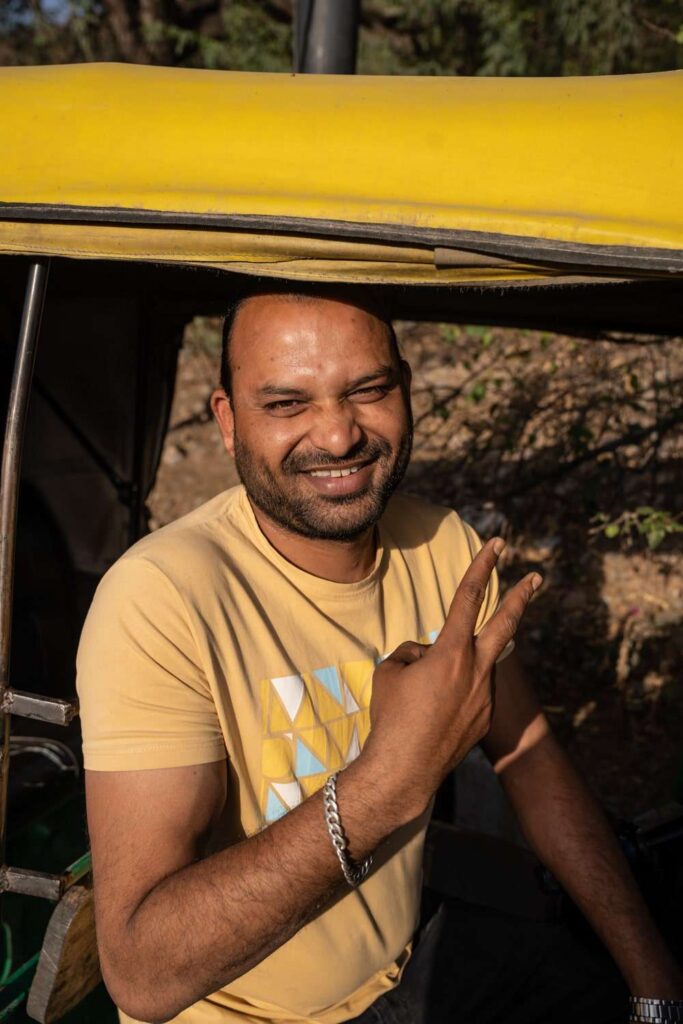 A man wearing a yellow t-shirt and silver bracelet sits in a yellow auto rickshaw, smiling and making a peace sign. Sunlight casts shadows on his face, capturing the carefree spirit of a 1-Week Rajasthan Itinerary among trees and foliage.