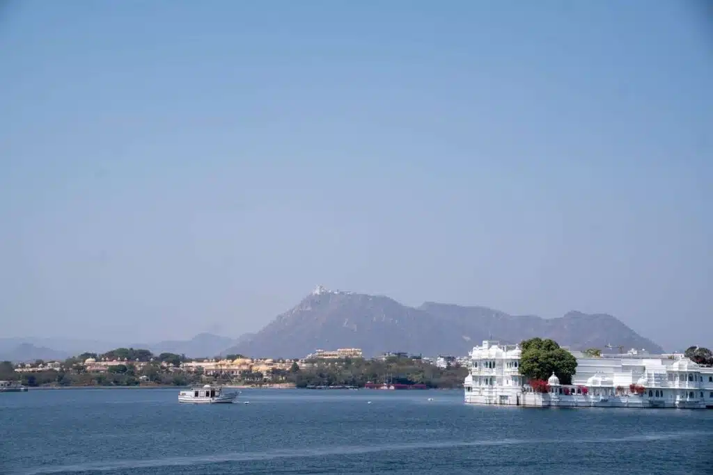 Backpacking India A serene lake with a white palace building on the water, a boat floating nearby, and distant mountains under a clear blue sky—an iconic moment on any 2-Week North India Itinerary.