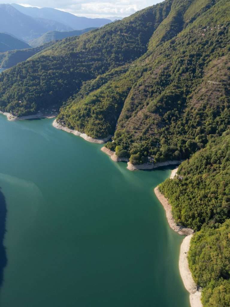 A scenic aerial view of Jablanica in Bosnia and Herzegovina, with a winding lake embraced by lush green hills. The calm water mirrors the vibrant foliage, while small sandy beaches line the shoreline. Distant mountain ranges rise under a clear blue sky.