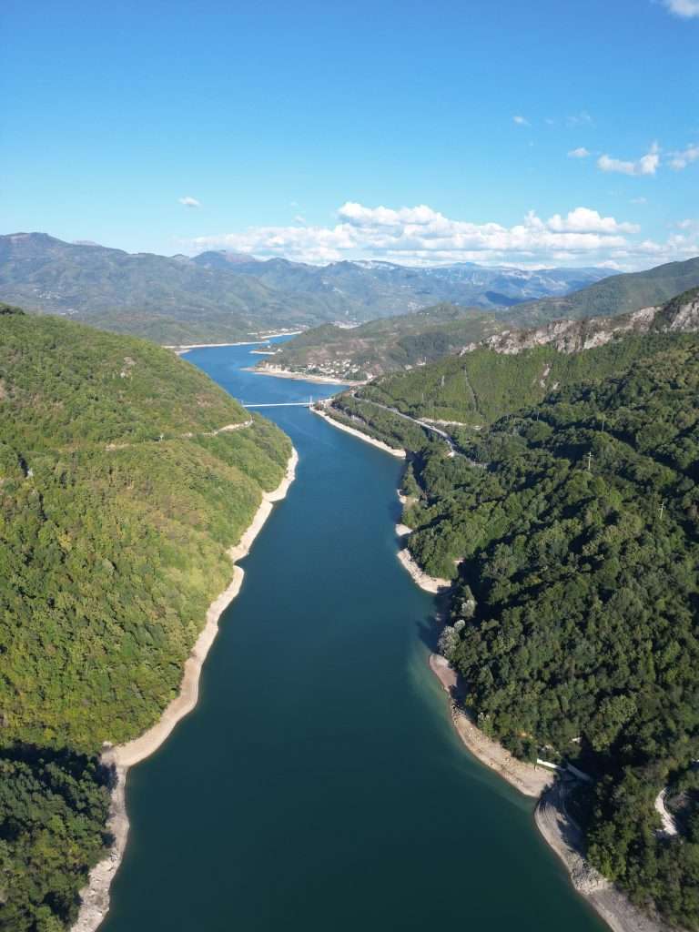 Aerial view of a winding river flowing through lush green mountains under a clear blue sky, reminiscent of 3 Days In Bosnia and Herzegovina. The dense forests cover the mountains, while the visible riverbanks lead the eye to distant hills and majestic horizons.