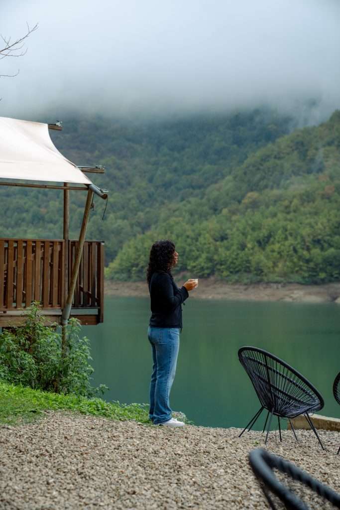 A person stands beside a serene lake enveloped by lush green hills and mist, as if savoring 3 Days in Bosnia and Herzegovina. They hold a cup, dressed casually. Nearby are black chairs and a wooden structure with a canopy, adding to the calm and peaceful atmosphere.