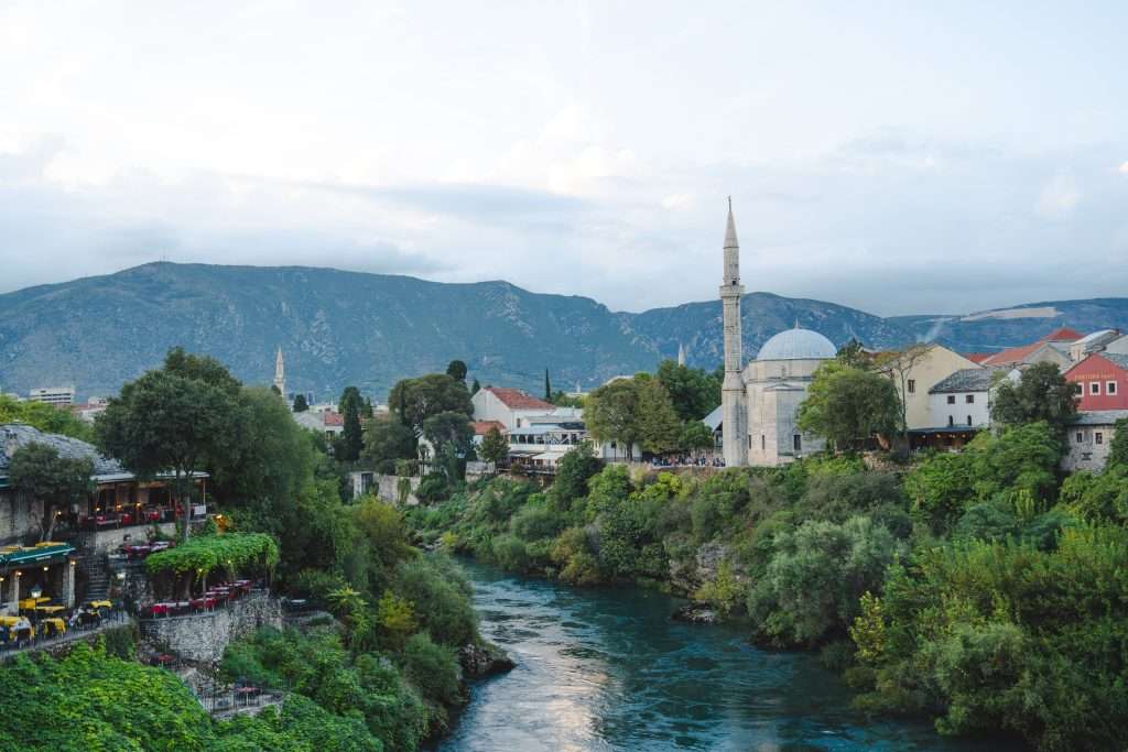 A scenic view unfolds as a river winds through Mostar's lush, green landscape. Historic buildings and a mosque with a tall minaret grace the right side, while rolling hills and cloudy skies serve as the backdrop. On the left, "One Day In Mostar" would not be complete without a café offering inviting outdoor seating.