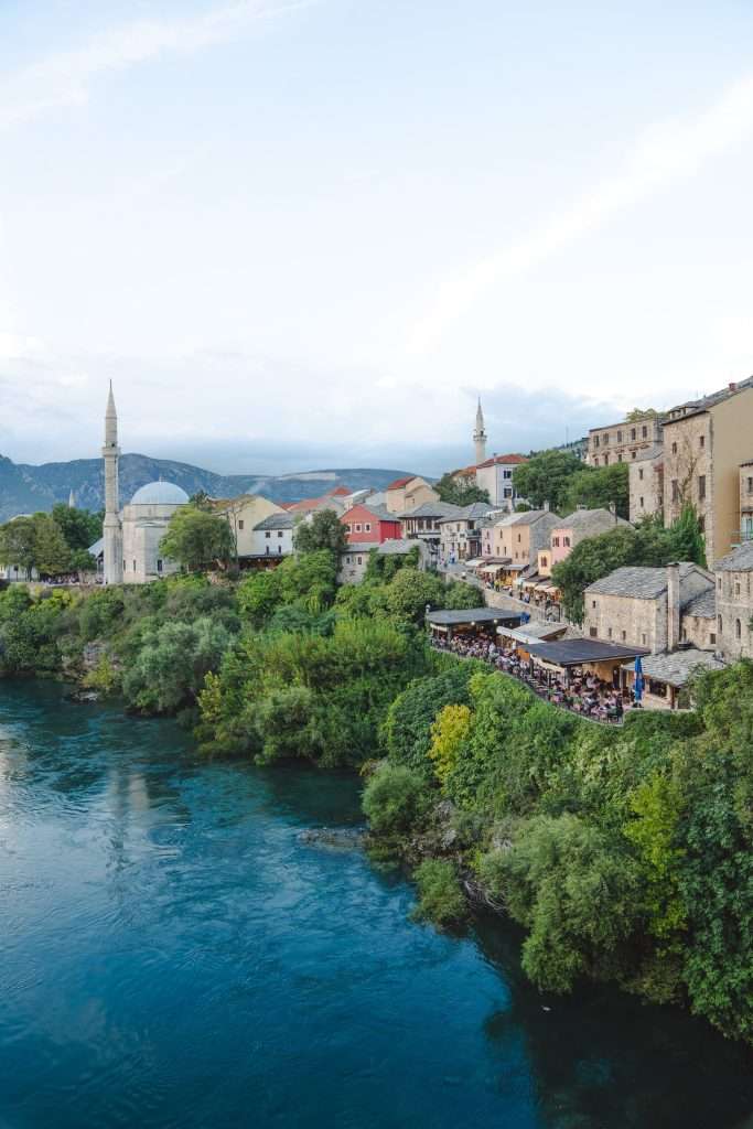 View of a picturesque riverside town with lush greenery and historic buildings. One day in Mostar, a calm river flows alongside, and a mosque with a tall minaret is visible. People are gathered in a riverside café, enjoying the scenic landscape.