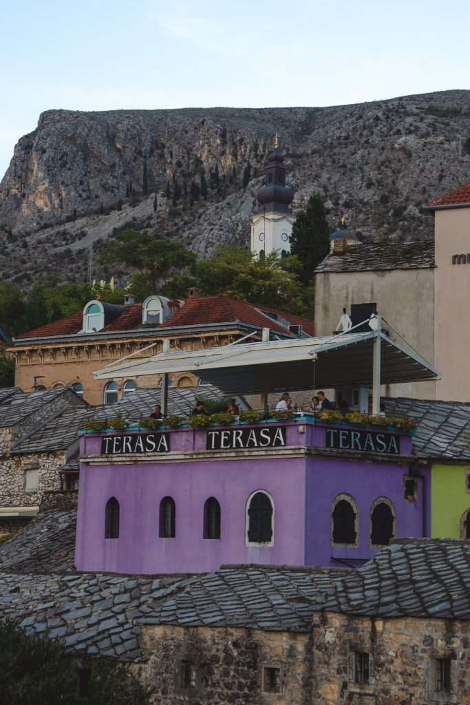 The purple building with a rooftop terrace labeled "TERASA" offers a perfect spot to enjoy the breathtaking views during One Day In Mostar. People take in the sights of the majestic mountain and iconic dome-spired structure under a partially cloudy sky, creating an unforgettable experience.