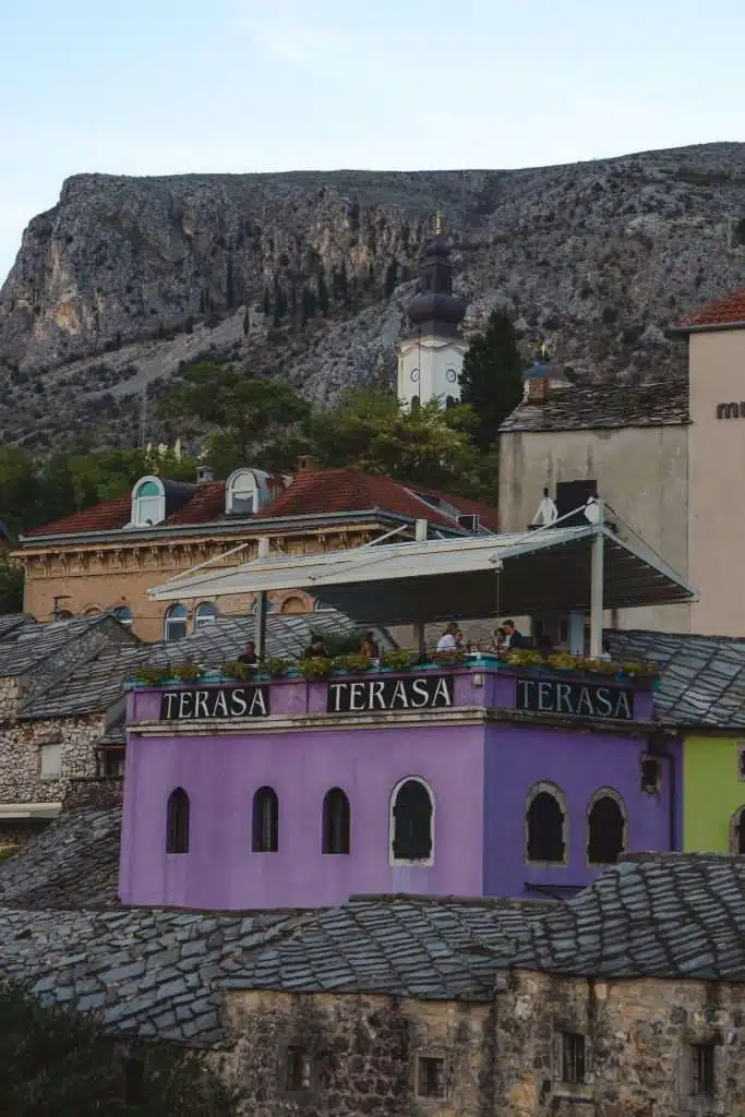 The purple building with a rooftop terrace labeled "TERASA" offers a perfect spot to enjoy the breathtaking views during One Day In Mostar. People take in the sights of the majestic mountain and iconic dome-spired structure under a partially cloudy sky, creating an unforgettable experience.