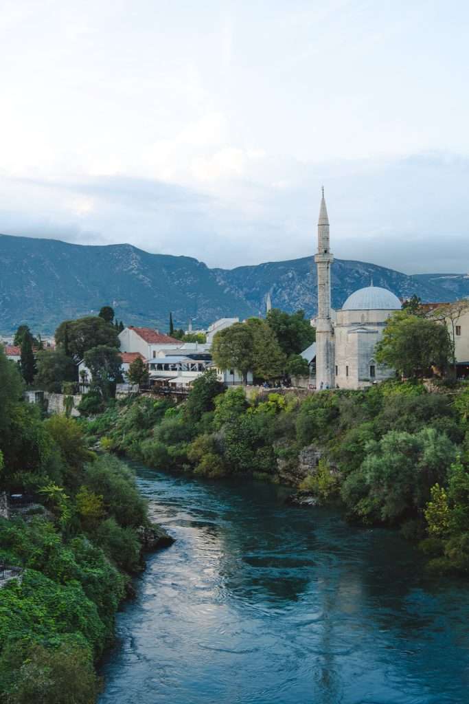 A scenic river winds through the lush landscape, graced by a mosque with a tall minaret and domed roof on its banks. In the background, mountains rise under a blue sky dotted with clouds—a view reminiscent of One Day In Mostar.