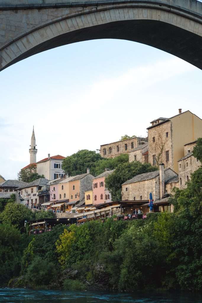 A scenic view of the historic town of Mostar, with stone buildings nestled under an arched bridge. A church spire and lush greenery are visible, complementing the river flowing below. The clear sky adds to the charm, making "One Day in Mostar" feel like a step back in time.