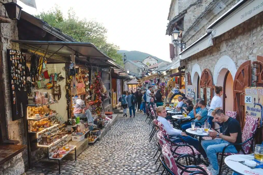 A bustling cobblestone street in Mostar, lined with shops and cafes. People are sitting at outdoor tables on the right, while souvenirs and colorful items are displayed for sale on the left. 
