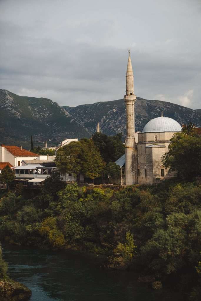 A scenic view of a mosque with a tall minaret and domed roof, surrounded by lush greenery and a river. Mountains rise in the background, under a cloudy sky, creating a serene landscape reminiscent of One Day in Mostar’s tranquil charm.