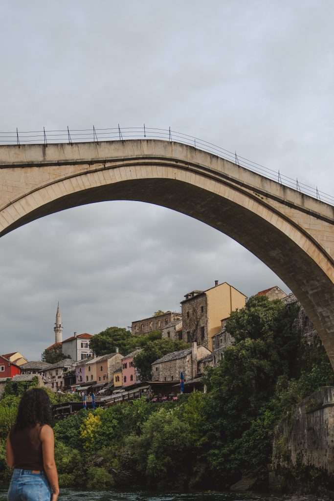 A person with curly hair stands gazing at Mostar’s large stone arch bridge. Behind it, the hillside is dotted with rustic buildings and lush greenery. The sky is overcast, and a minaret rises in the background—a perfect scene for one day in Mostar.