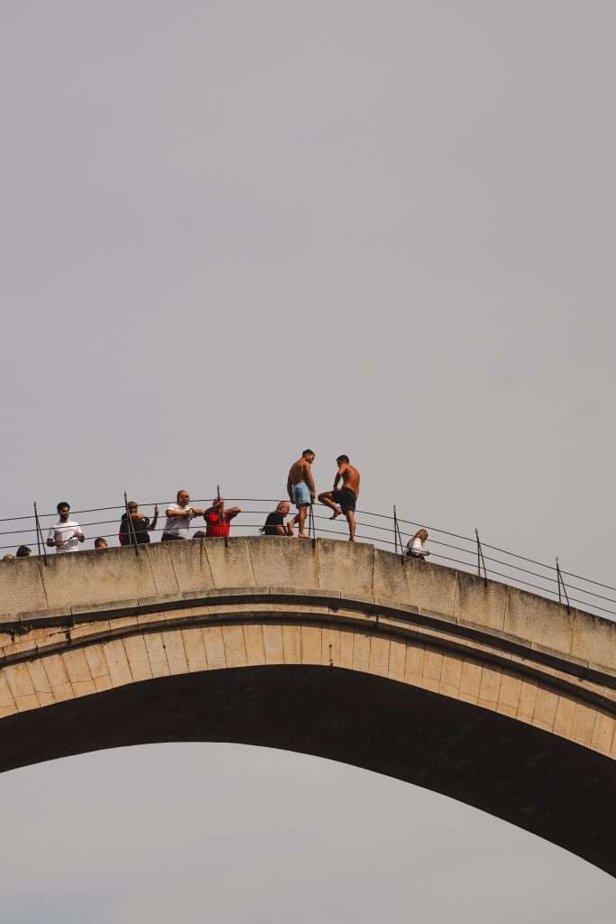 During One Day In Mostar, people gather on a stone arch bridge, with a few individuals preparing to jump off while others watch from the railings beneath the clear sky.