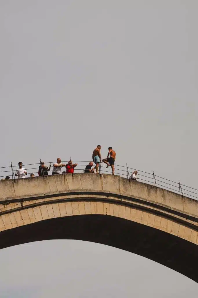 People gather on a stone arch bridge in Mostar, with a few individuals preparing to jump off while others watch from the railings beneath the clear sky.