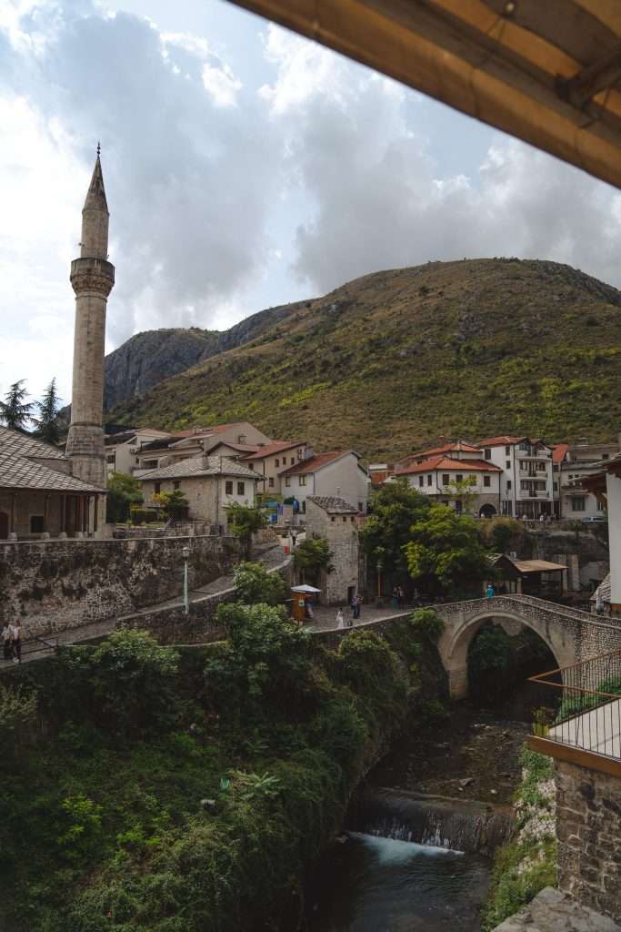 Experience the scenic view of a historic town, reminiscent of one day in Mostar, with a stone bridge arching elegantly over a river. Traditional houses and a tall minaret complement the landscape, while a hillside rises proudly in the background under a partly cloudy sky.