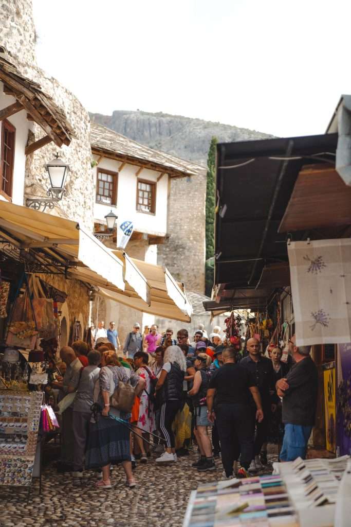 A bustling outdoor market scene, reminiscent of "One Day In Mostar," features people shopping along narrow stone-paved streets lined with stalls offering various items. Traditional buildings with wooden windows encircle the lively area, creating a vibrant hub filled with a diverse crowd.