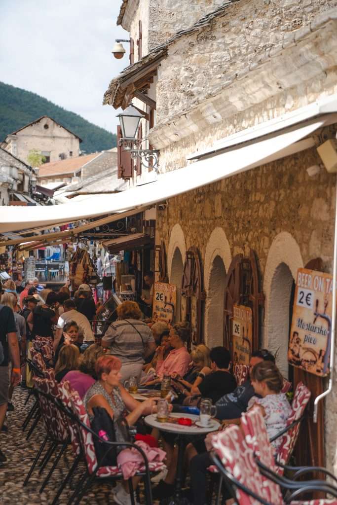 A lively cobblestone street in Mostar is lined with outdoor cafes and shops. People are seated, eating, and conversing under a canopy. The sign advertises beer on tap for 2€. Historic stone buildings and hills form the backdrop of this picturesque scene for those spending one day in Mostar.